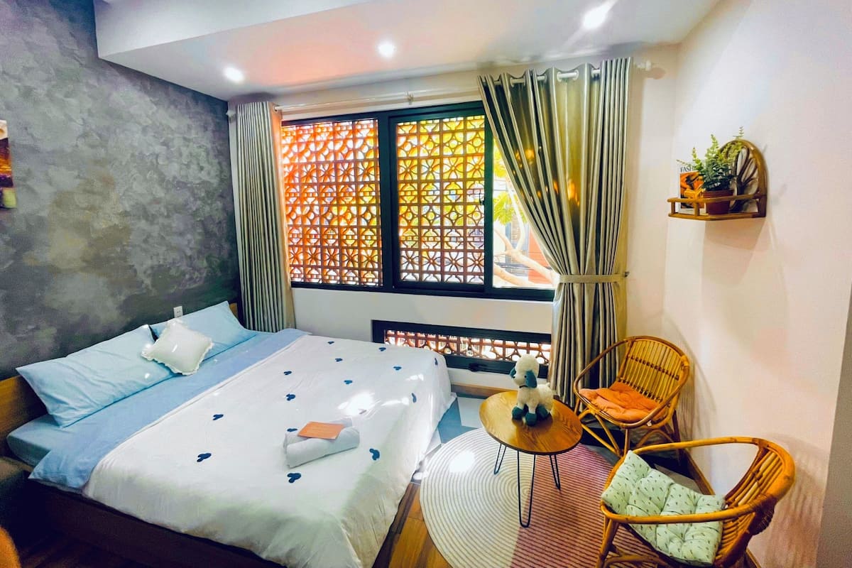 Une chambre d’hôtel avec un lit, une petite table ronde, deux chaises en osier, une fenêtre avec des rideaux et une étagère avec une plante.