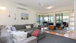 A living room with a grey sectional sofa, a wooden coffee table, and a ceiling fan. There is a large window providing a view of the outdoors.