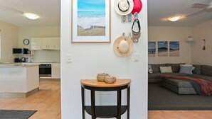 A modern kitchen with white cabinets, a microwave, and a coffee maker. There is a dining area with a round wooden table and a black chair. The living room features a gray sofa and a red blanket.