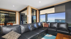 A modern living room with a black leather sectional sofa, a wooden coffee table, and large windows offering a view of the sea.