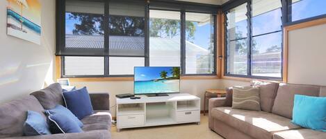 Ein Wohnzimmer mit einem großen Fernseher, zwei Sofas und einem Fenster mit Blick auf einen Strand.