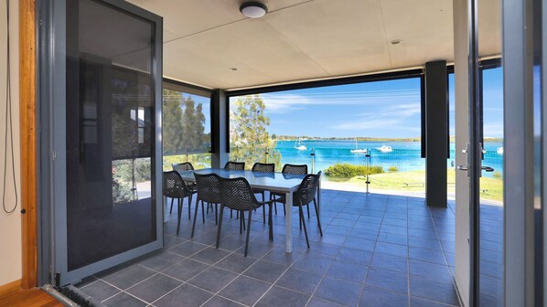 A modern dining area with a table and chairs, a glass door leading to a balcony with a view of the sea and boats.