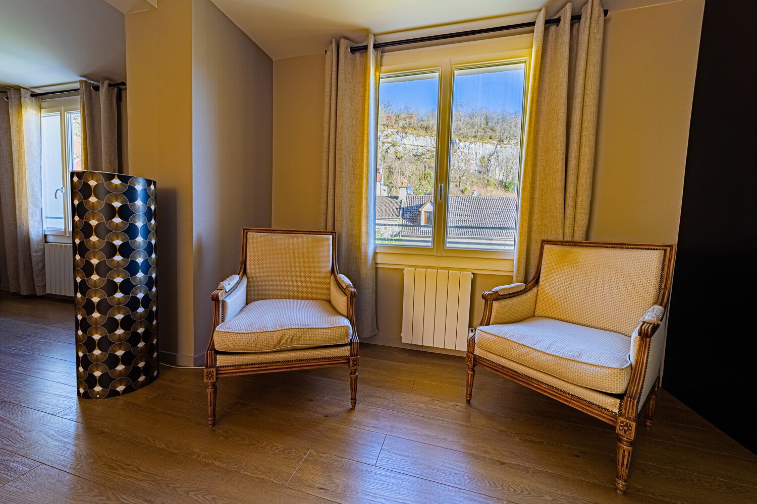 Una habitación con dos sillones de mimbre, un pilar decorado y un ventanal con vistas a edificios y árboles.