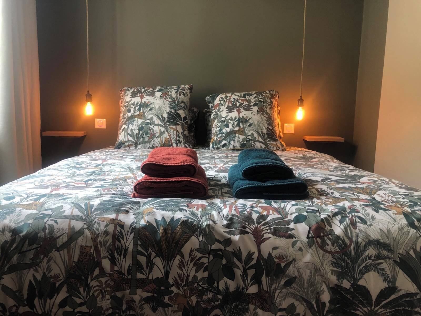 Un lit recouvert d’une couvre-lit à motifs floraux, deux oreillers à motifs de feuilles et deux serviettes pliées, une rouge et une bleue, posées sur le lit.