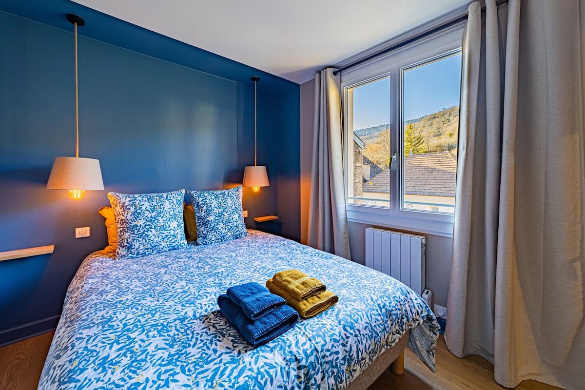 A bedroom with a large bed, two bedside lamps, a window with curtains, and a view of a hilly landscape.