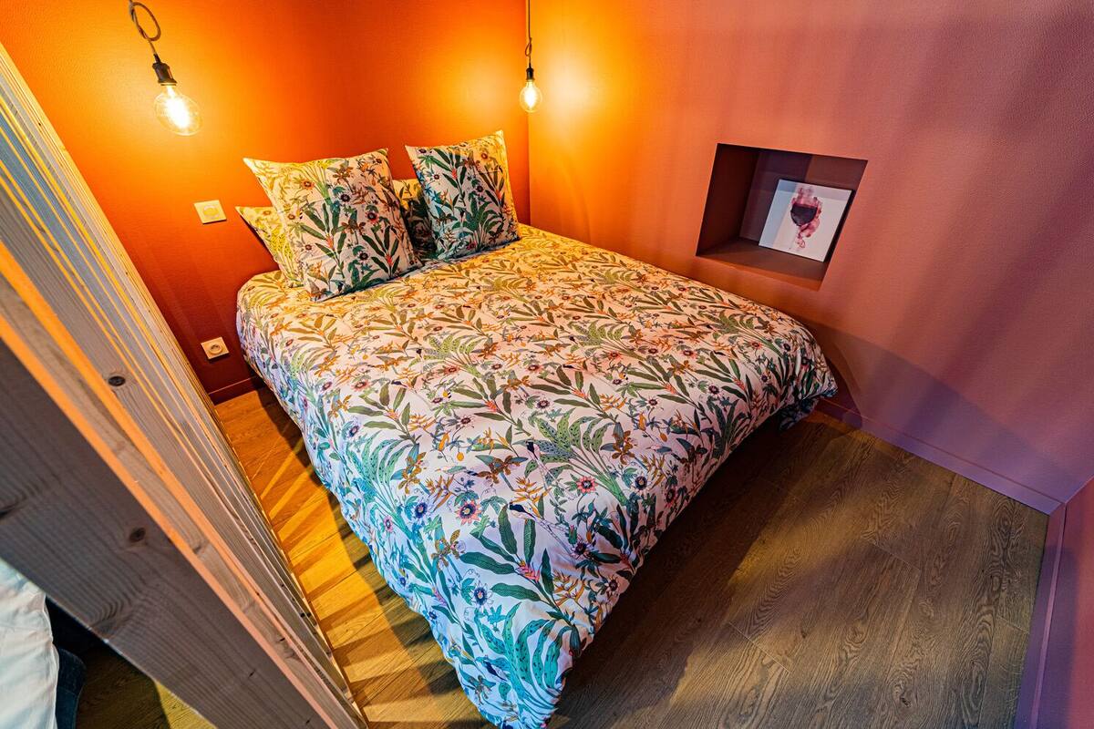 A bedroom with a floral bedspread, two wall-mounted light fixtures, a small framed picture, and a wooden floor.