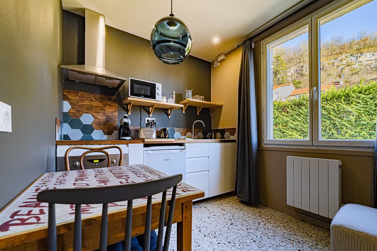 A compact kitchen with a dining table, chairs, a hanging light fixture, and a view of the outdoors through a window.