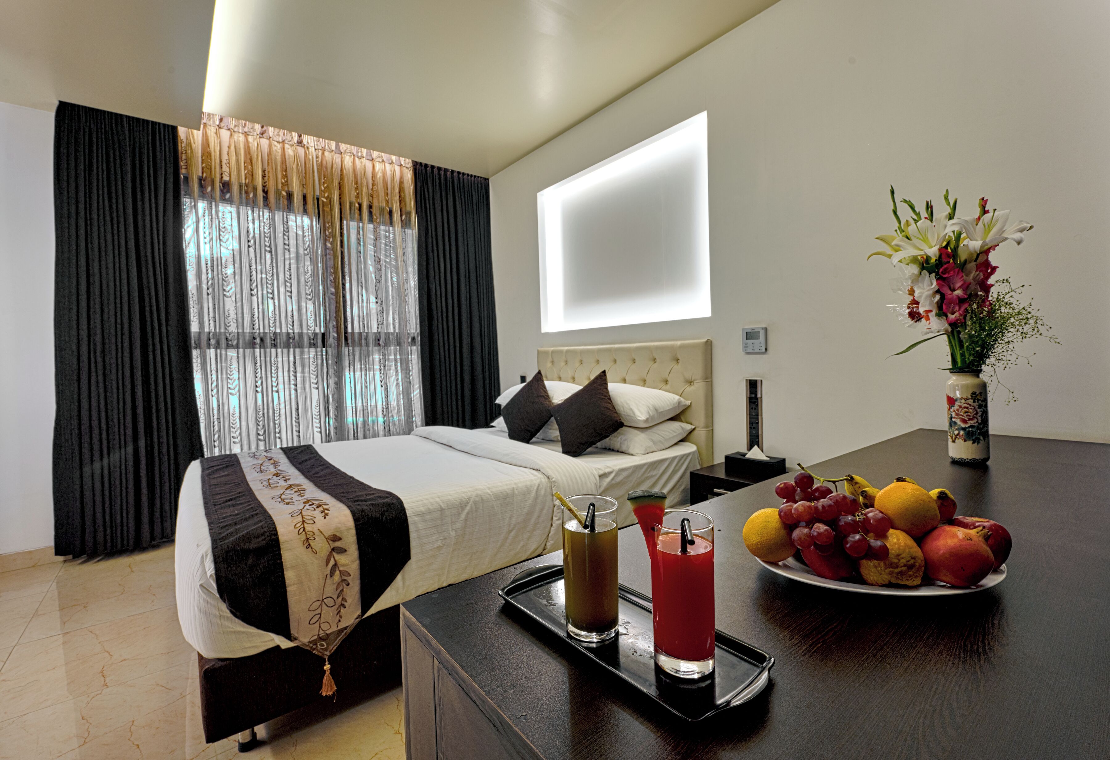 A hotel room with a bed, bedside table with drinks, a fruit plate, and a vase with flowers.