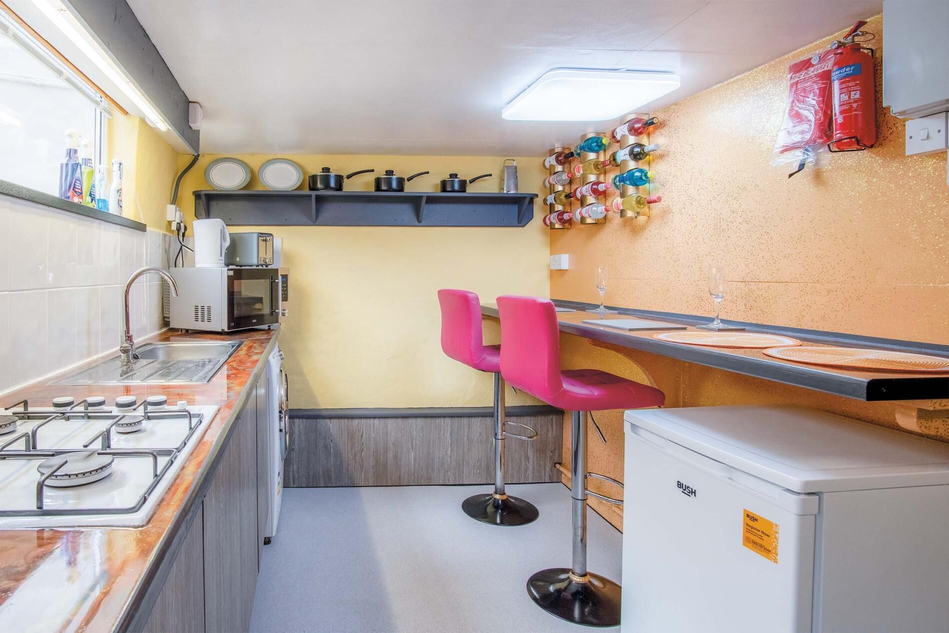 A compact kitchen with a gas stove, microwave, and toaster. There are two pink bar stools and a small refrigerator.