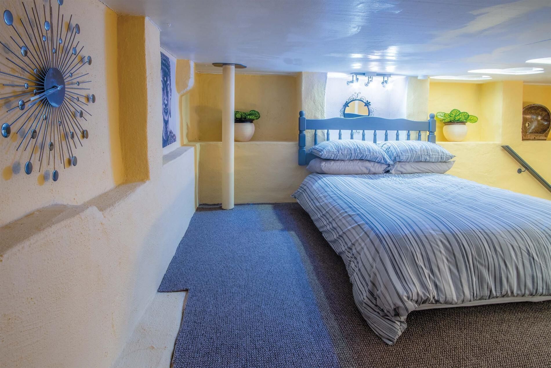 A bedroom with a bed, a blue headboard, a wall clock, and a potted plant.