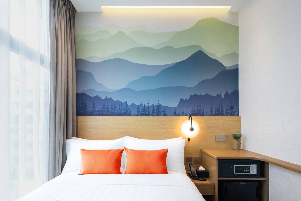 Ein Hotelzimmer mit einem Bett, einem Nachttisch, einer Lampe und einer Wandmalerei von Bergen.