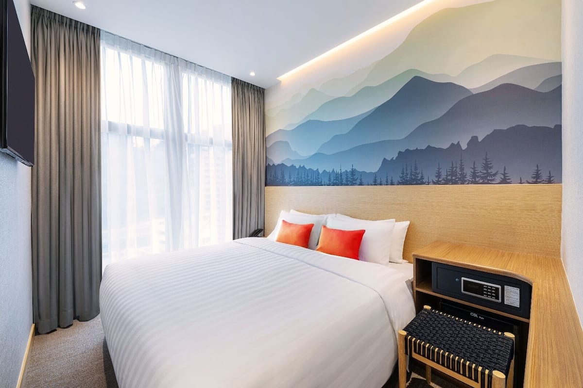 Ein Hotelzimmer mit einem großen Bett, einem Kopfteil mit einem Gebirge als Gemälde, einem Schreibtisch mit Tastatur und einem Fenster mit Vorhängen.