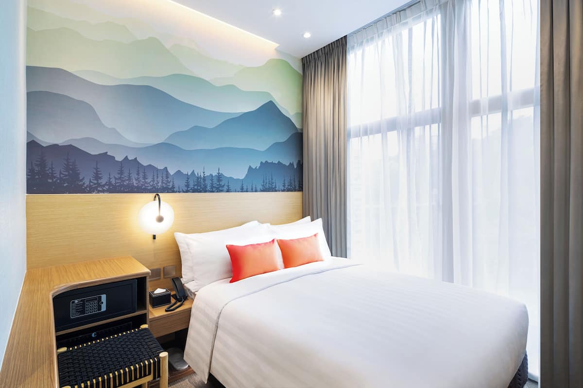 Ein Hotelzimmer mit Bett, Nachttisch mit Lampe, Tastatur und Wandgemälde von Bergen.