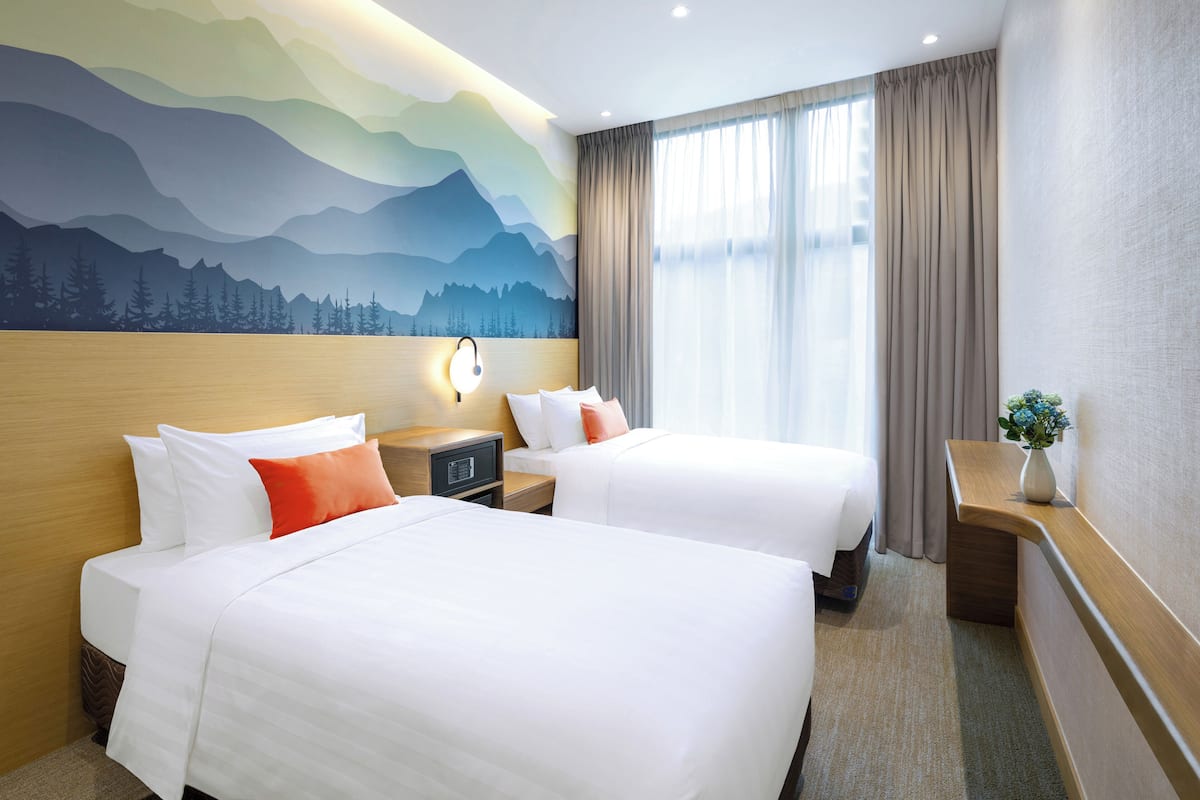 Ein Hotelzimmer mit zwei Betten, einem Nachttisch, einer Lampe, einem Fenster mit Vorhängen und einem Wandbild.