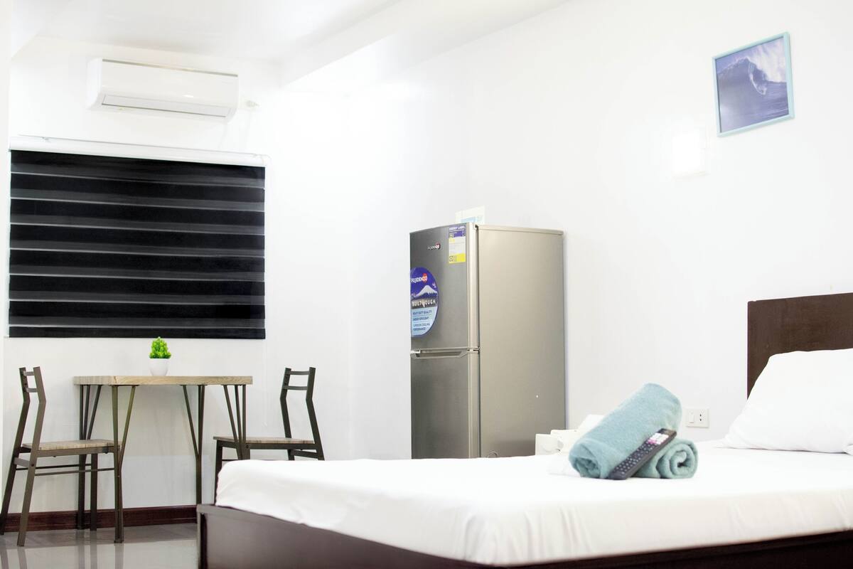 A hotel room with a bed, a small table with chairs, a refrigerator, and a window with blinds.