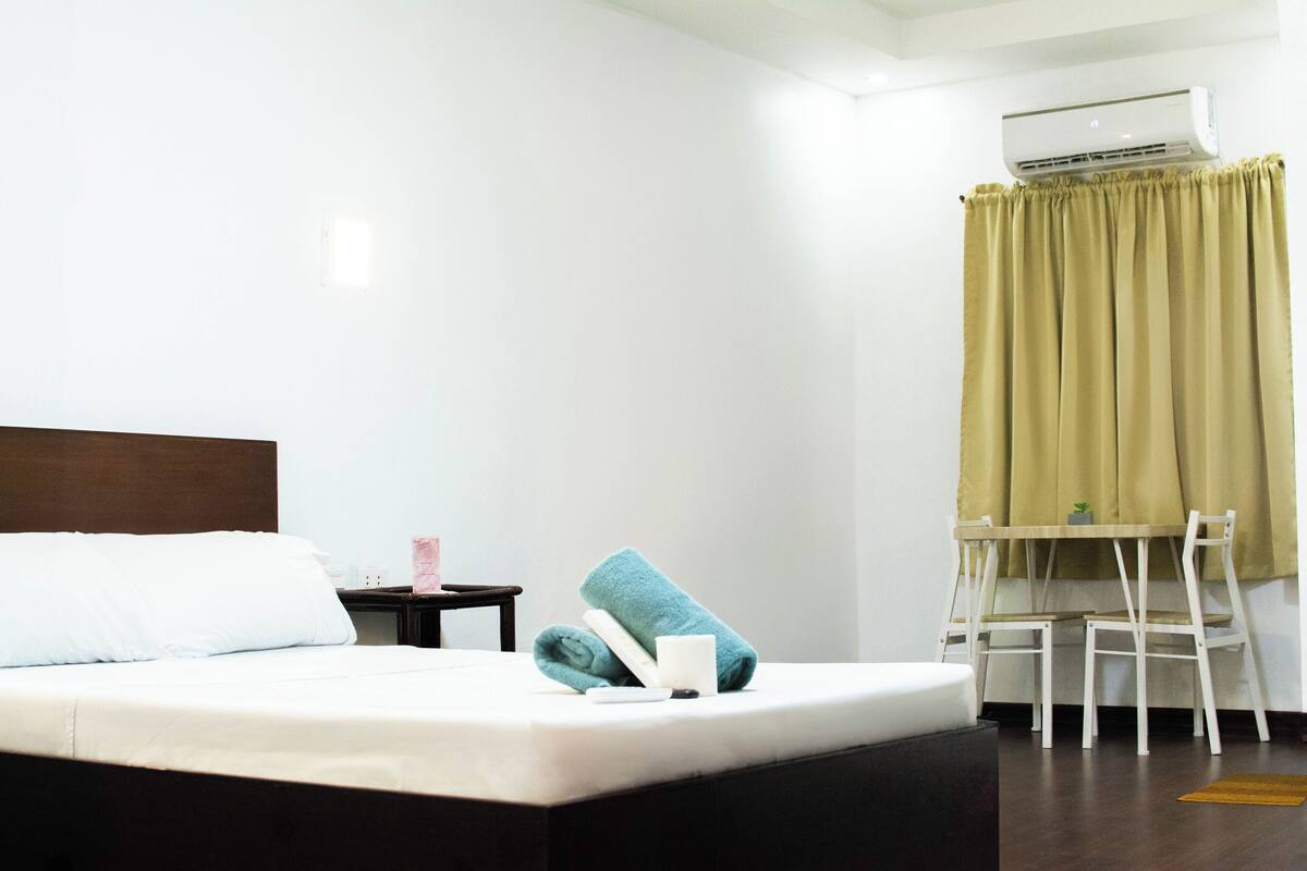 A hotel room with a bed, a desk, a chair, an air conditioner, and a window with curtains.