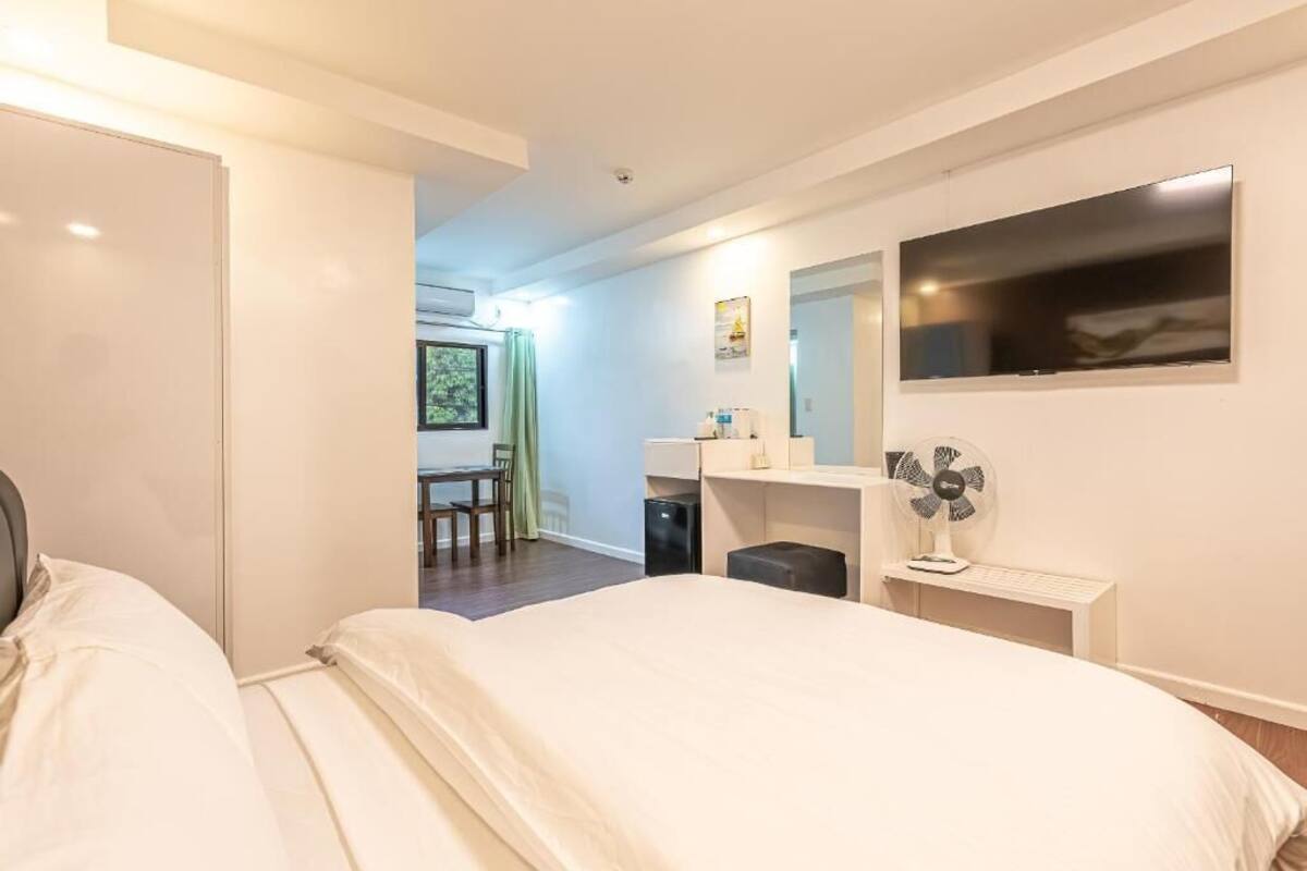 A modern hotel room with a large bed, a desk, a chair, a small table, a mirror, and a TV.