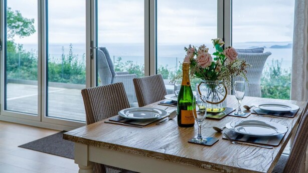 A dining table set with plates, cutlery, and a bottle of Veuve Clicquot champagne, with a view of the sea through large windows. - Shearwater 30 - 2 Bedroom Lodge - Summerhill (Narberth)