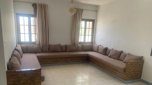 A large L-shaped sectional sofa with a wooden frame and a patterned border, positioned in a room with tiled flooring and two windows with curtains. - Villa Soliman Plage s+3 (Nabeul)