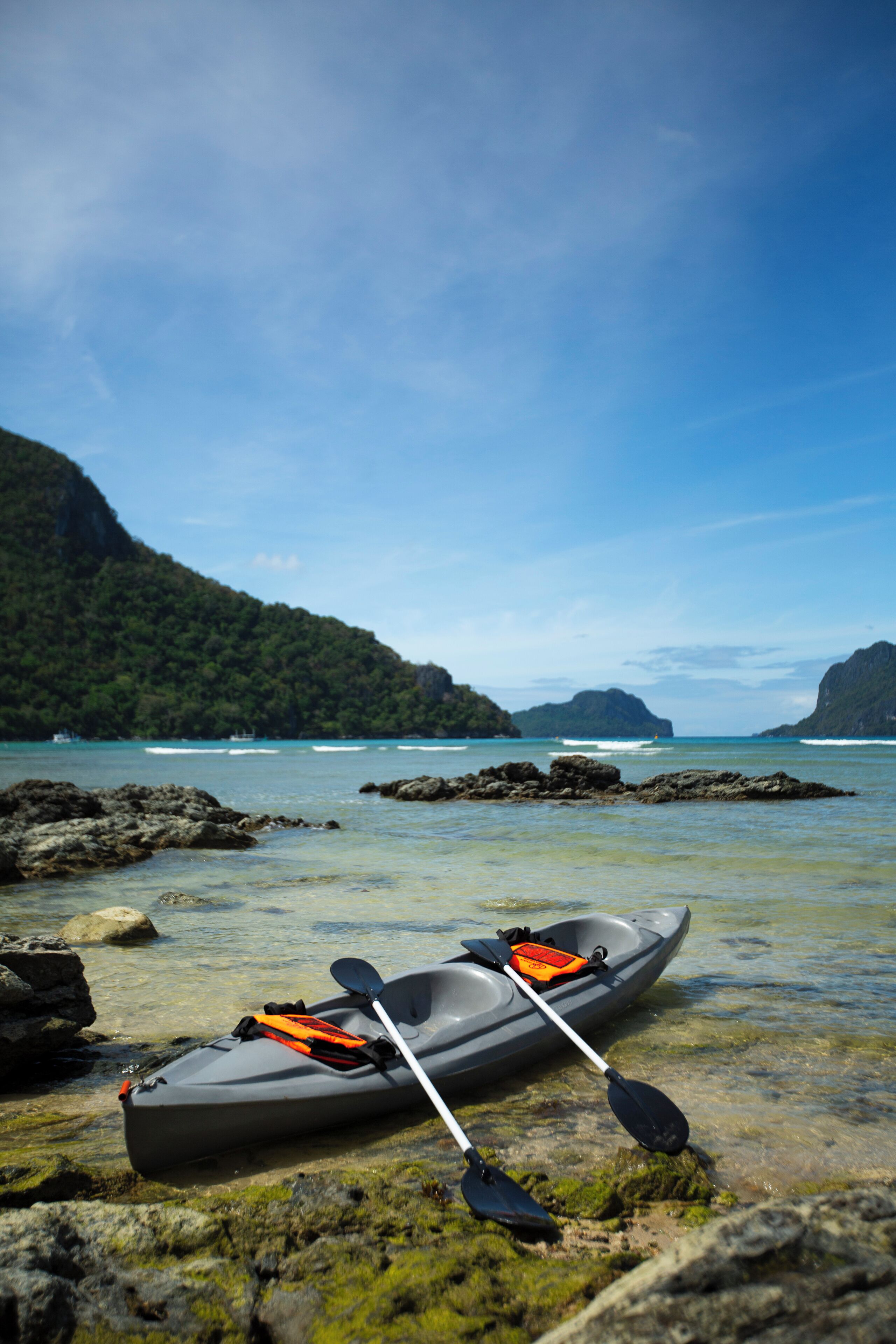 A kayak with paddles and an orange life jacket on a rocky shore with clear water and distant mountains.