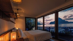 Un dormitorio con un ventanal grande que da a una masa de agua y montañas, una cama con ropa blanca y una lámpara de pared con textura.