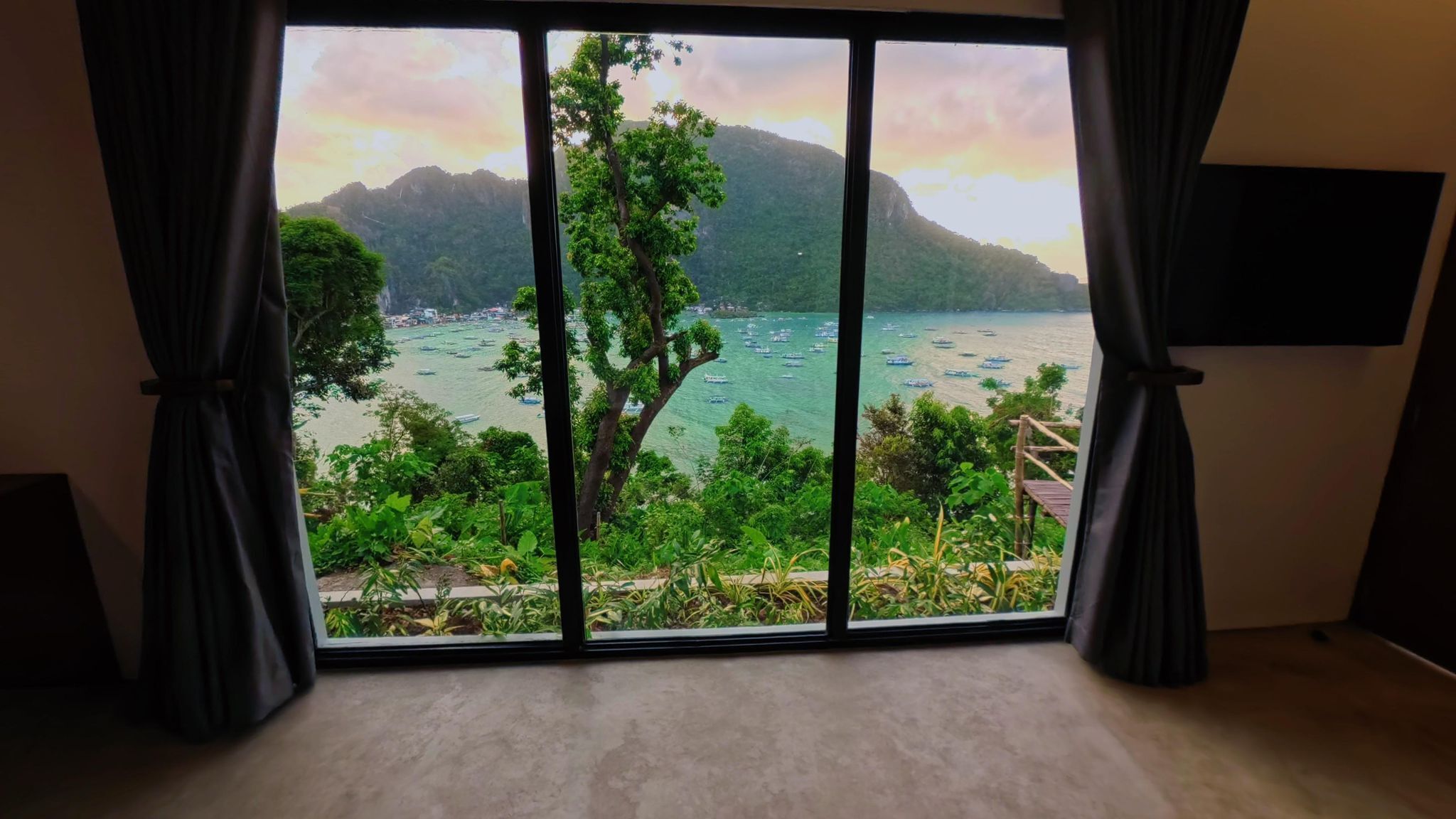 Una habitación con un amplio ventanal que ofrece vistas a una zona costera con barcos, vegetación y montañas.