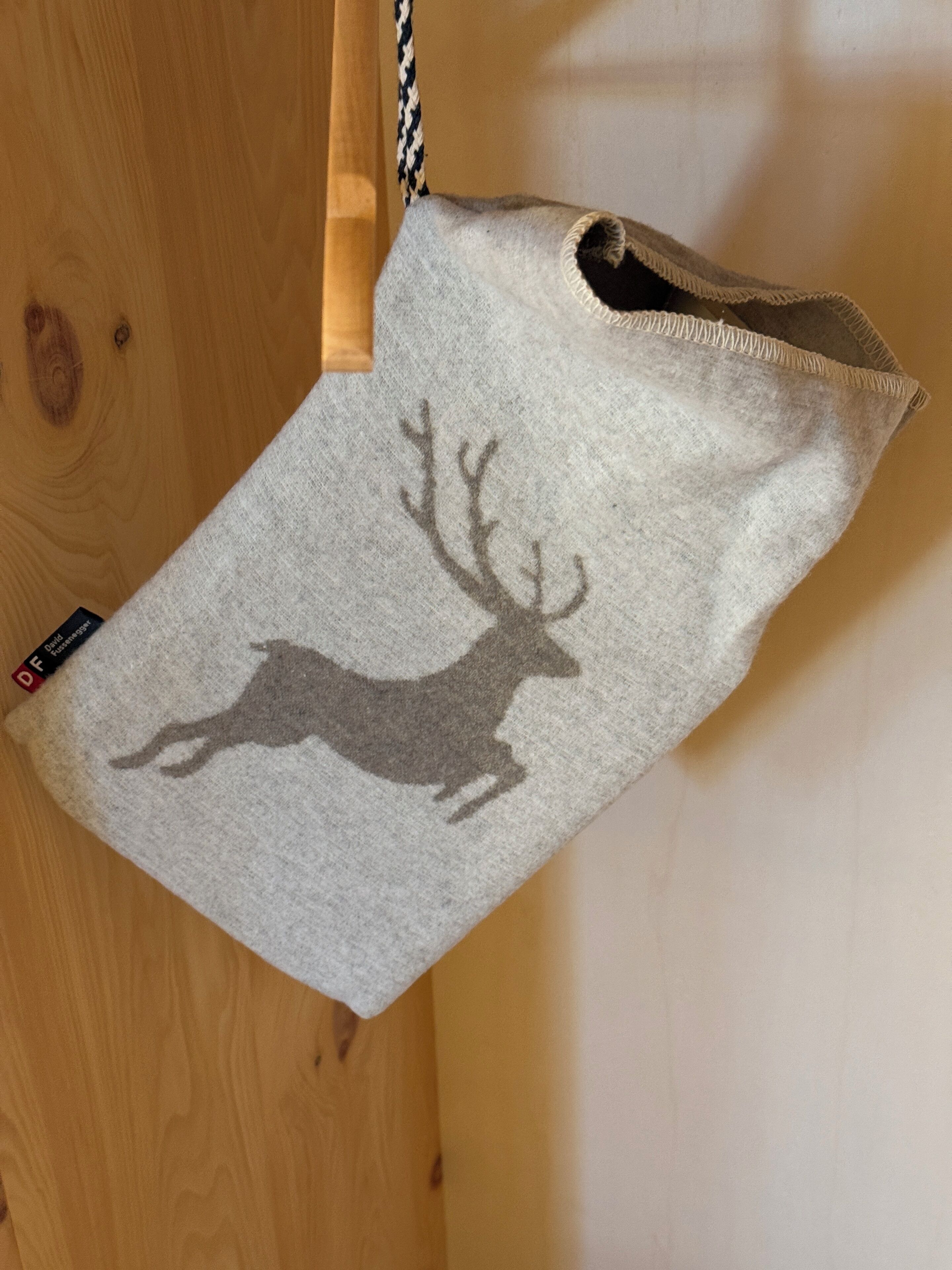 Une pochette grise en feutre ornée de la silhouette d’un cerf, suspendue à une porte en bois.