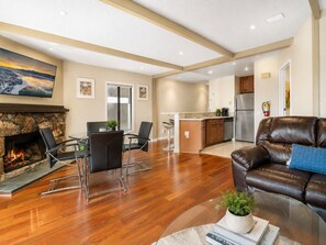 A modern living room with a fireplace, a leather sofa, a dining area with chairs, and a kitchen in the background.