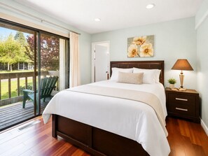 A bedroom with a large bed, a nightstand with a lamp, a painting on the wall, and a view of a deck and trees. - Lakeview Condo - Lakefront Complex, Lake Views, Heated Pool (Big Bear Lake)