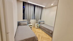 Um quarto pequeno e moderno com duas camas de solteiro, uma mesa de cabeceira com abajur e uma janela com cortinas leves.