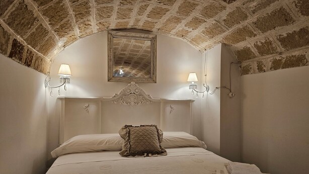 A bedroom with a brick ceiling, a bed with a quilted pillow, two wall-mounted lamps, and a mirror above the bed. - Le Camerette in Monopoli (Monopoli)