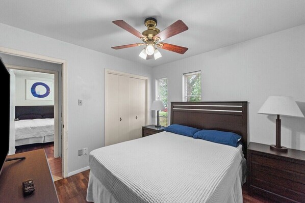 Un dormitorio con cama, ventilador de techo, mesita de noche, lámpara y puerta que da a otra habitación.
