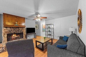 A living room with a stone fireplace, a ceiling fan, a grey sofa, and a television.
