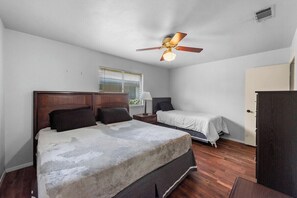 Une chambre avec un grand lit, un plus petit lit, un ventilateur de plafond, une fenêtre avec des stores et une porte.
