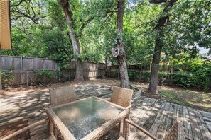 Eine Terrasse im Freien mit Korbmöbeln und einem Glastisch, umgeben von Bäumen und einer Holzterrasse.
