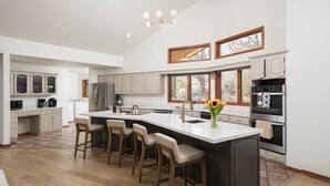 A spacious kitchen with a large island, bar stools, and a view of the outdoors through large windows. - Red Rock Vista | Incredible Views, Hot Tub, Firepit, Trails (Sedona)
