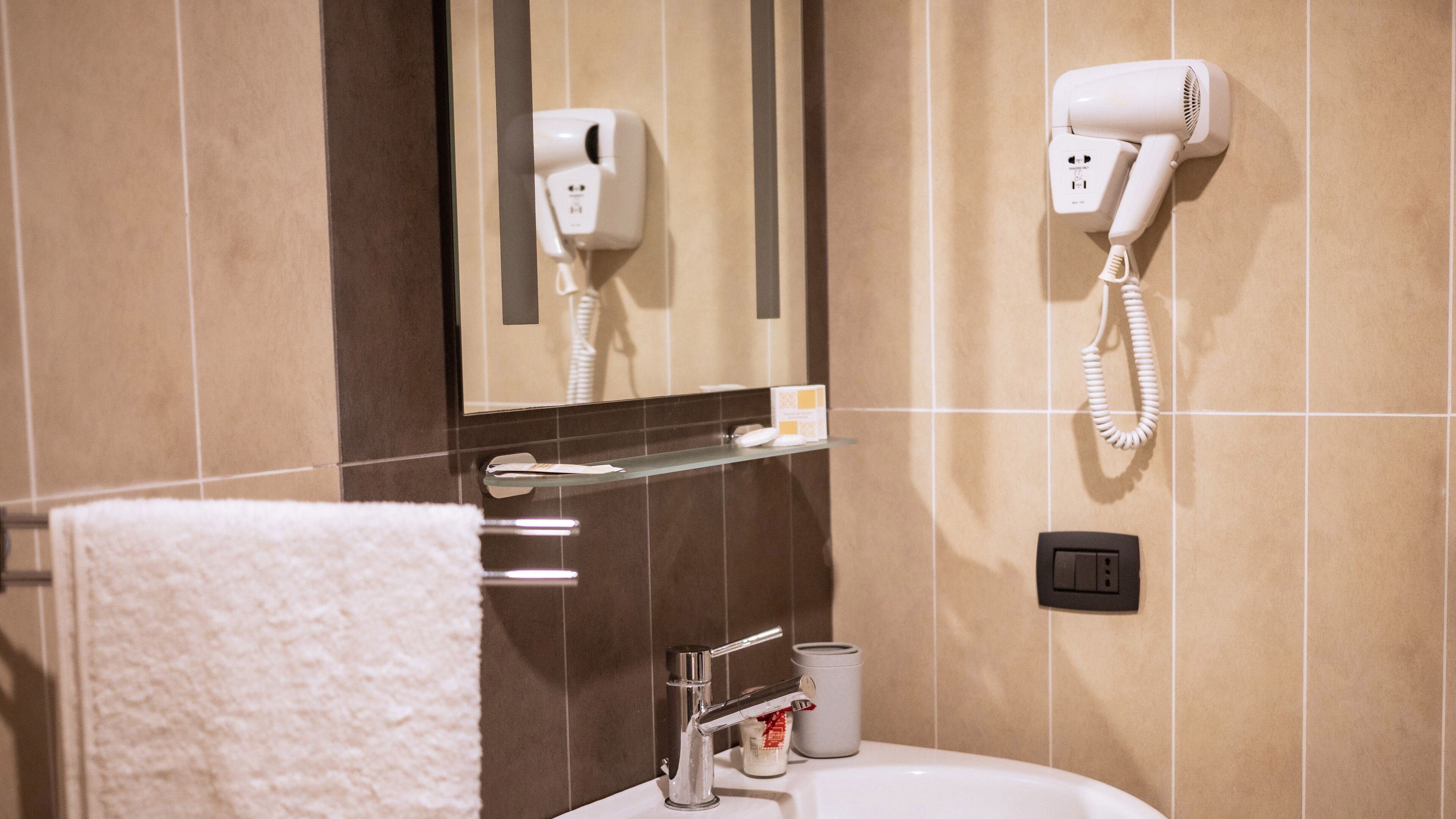 Une salle de bain avec un lavabo blanc, un porte-serviettes avec une serviette blanche, un miroir, un sèche-cheveux et un distributeur de savon.
