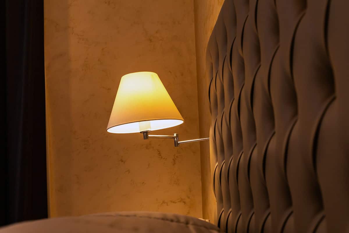 A wall-mounted lamp with a conical shade, a textured wall, and a cushioned headboard.