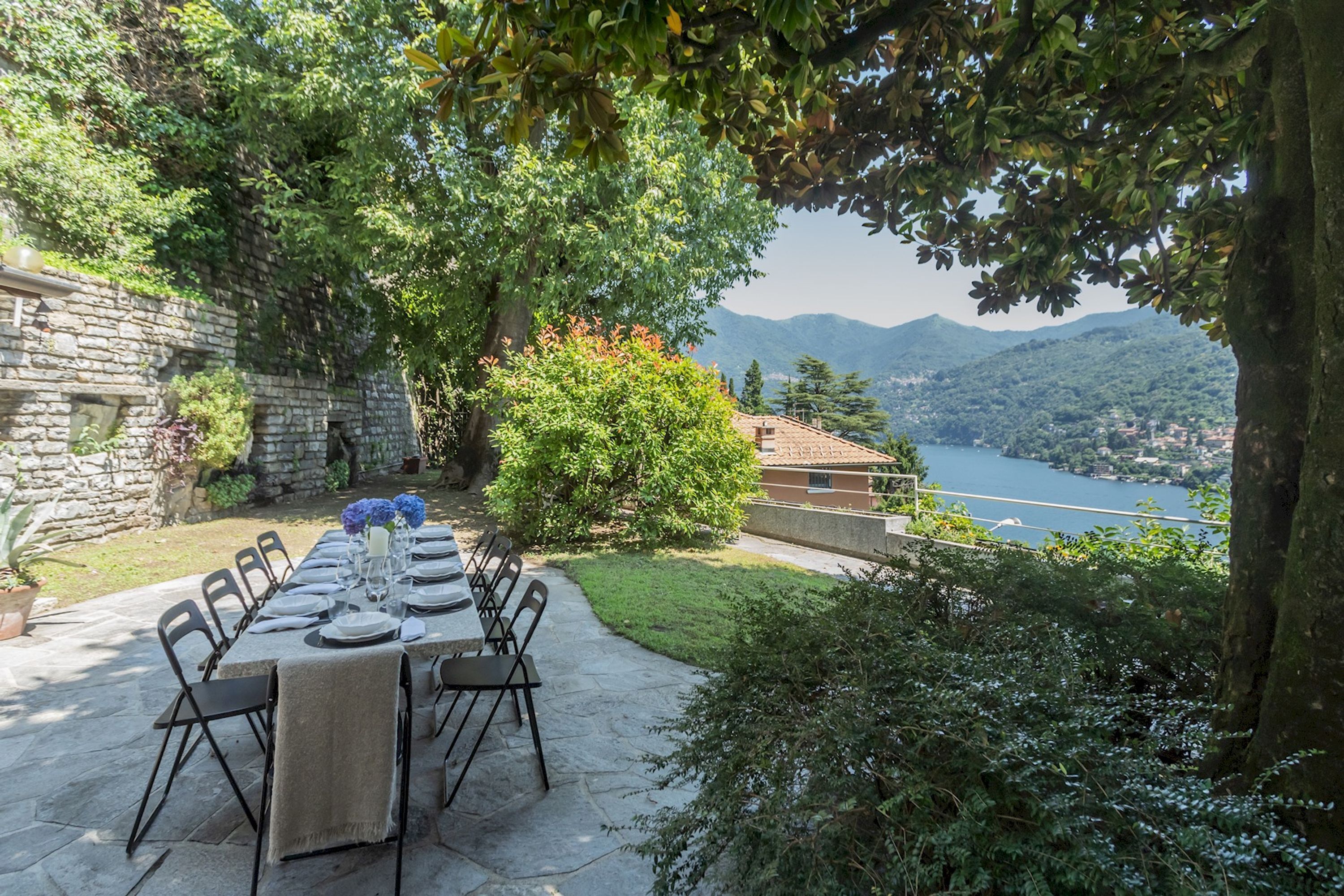 A stone table set for dining with chairs, a view of a lake and mountains, and a stone wall.