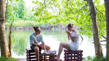 Two people sitting on wooden benches by a calm lake, with a small table between them holding cups and a teapot.
