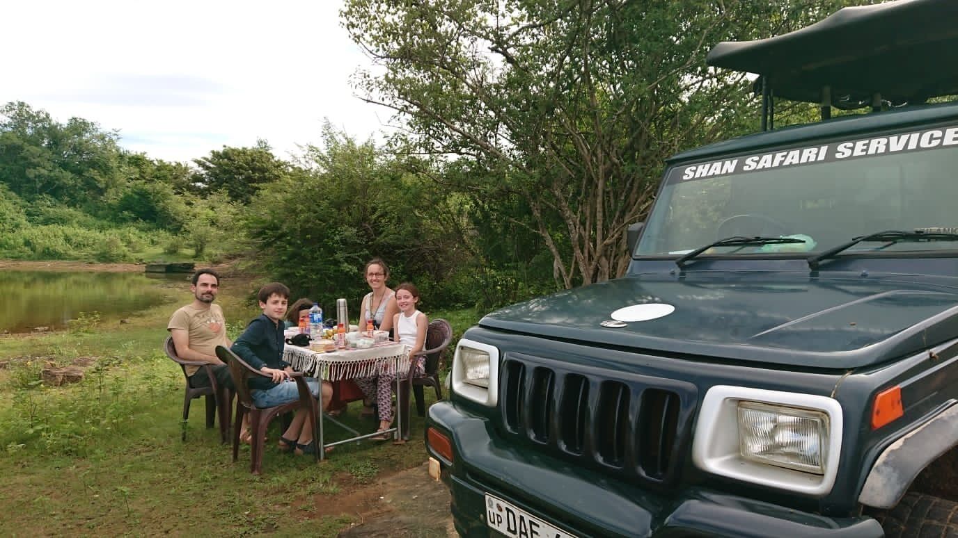 A group of people sitting at a table outdoors with a vehicle labeled "SHAN SAFARI SERVICE" in the foreground.