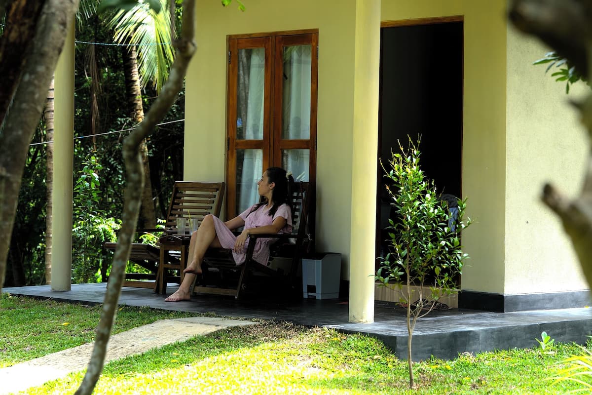 A single-story building with a porch, a small tree, and a person sitting on a chair.
