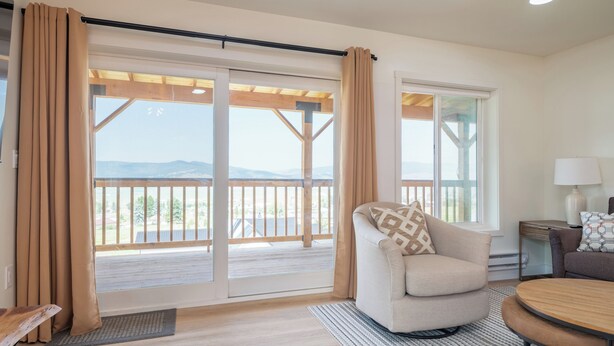 Ein modernes Wohnzimmer mit einer Couch, einem Couchtisch und Blick auf einen Balkon mit einer Holzdielenveranda und Bergen im Hintergrund.