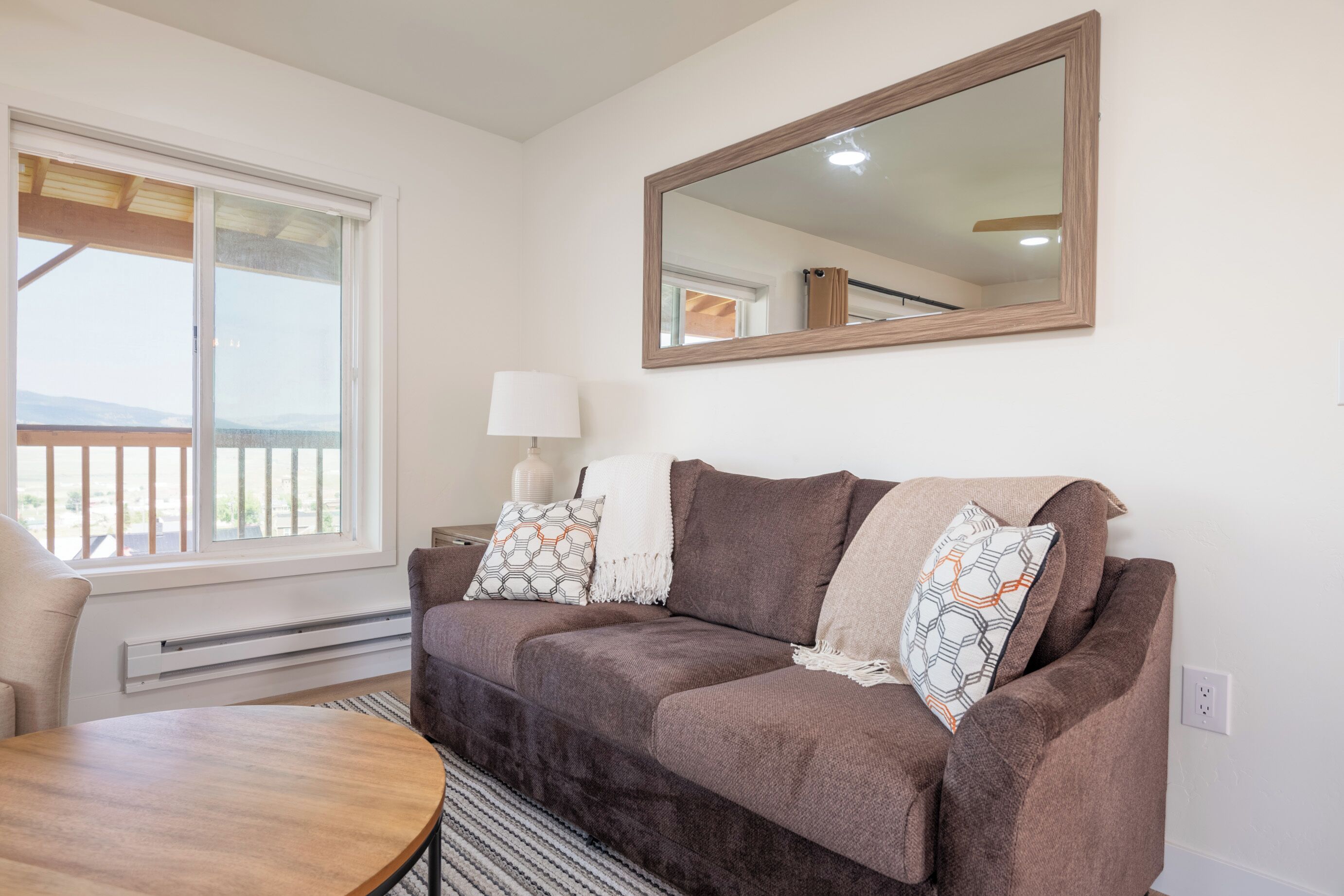 A living room with a brown sofa, a round wooden table, and a large mirror on the wall.
