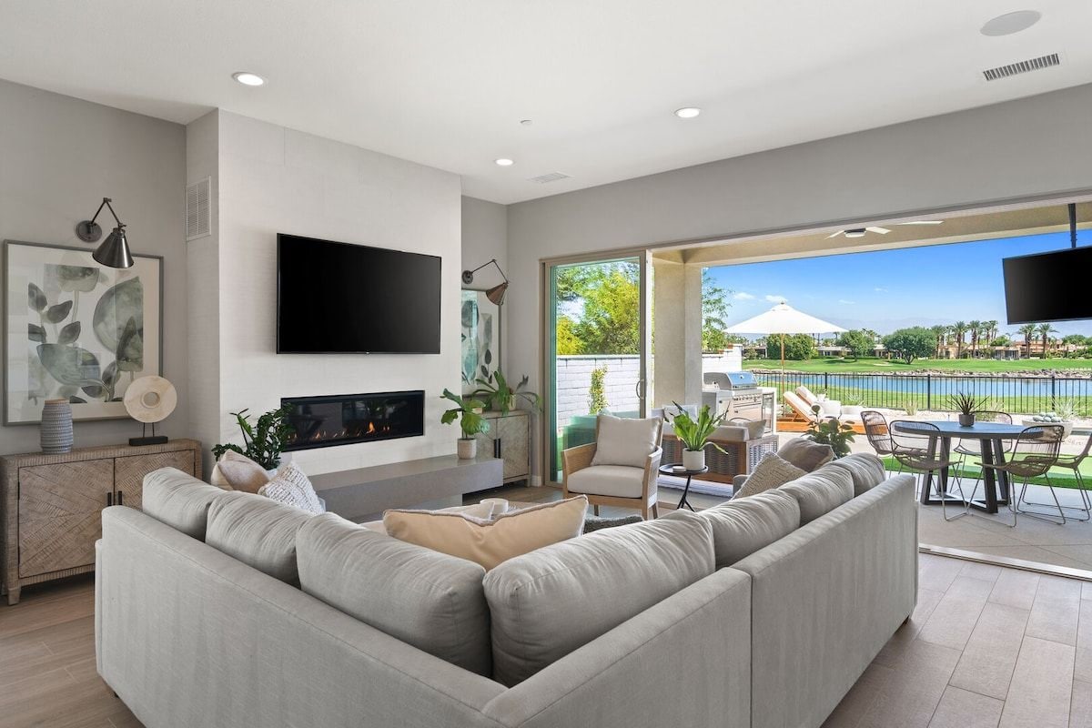 A modern living room with a flat-screen TV, a fireplace, and a sofa. There is a dining area with outdoor seating and a view of a pool.