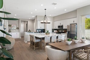 A modern kitchen with white cabinetry, a central island with bar stools, and a dining area with a wooden table. - Saltwater Pool Haven, EV Charger, and Golf Views! (La Quinta)
