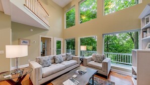 A spacious living room with a high ceiling, large windows, a staircase, and a view of greenery outside. - Alpine Chalet| 5BR | Helen, GA| near golf| community pool (Helen)