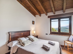 A bedroom with a wooden ceiling, a large bed with pillows, a nightstand, a window with a view, and a small table with a chair.