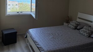 A bedroom with a bed, a nightstand, a small stool, a window with a view of the sea, and a ceiling light. - Holiday Home 'Chez Cisco' with Sea View, Wi-Fi and Air Conditioning (le lavandou)