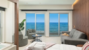 A modern hotel room with a large sliding glass door leading to a balcony with a view of the sea. There is a bed, a gray sofa, and a small round table with a plant. - Elia Sea Suites (Chania)